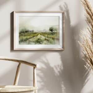 Op de afbeelding: Een aquarel van een wijngaardlandschap met een pad dat door de rijen wijnstokken loopt. Het schilderij is ingelijst in een licht houten lijst en hangt aan een lichtbeige muur.