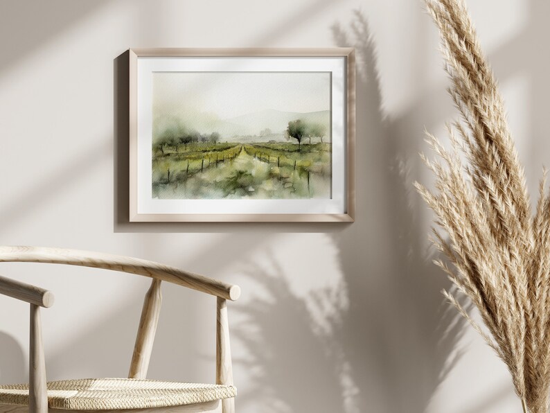 Op de afbeelding: Een aquarel van een wijngaardlandschap met een pad dat door de rijen wijnstokken loopt. Het schilderij is ingelijst in een licht houten lijst en hangt aan een lichtbeige muur.