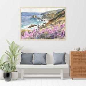 May include: Watercolor painting of a coastal scene with a rocky cliff, ocean waves, and pink wildflowers. The painting is framed and hanging on a white wall above a white bench with gray cushions.
