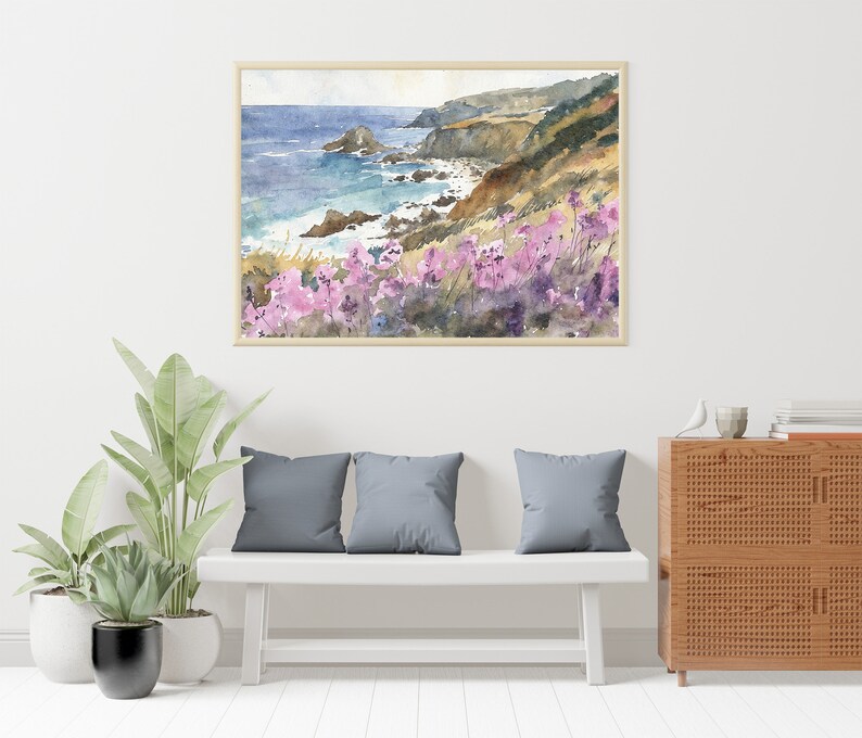 May include: Watercolor painting of a coastal scene with a rocky cliff, ocean waves, and pink wildflowers. The painting is framed and hanging on a white wall above a white bench with gray cushions.