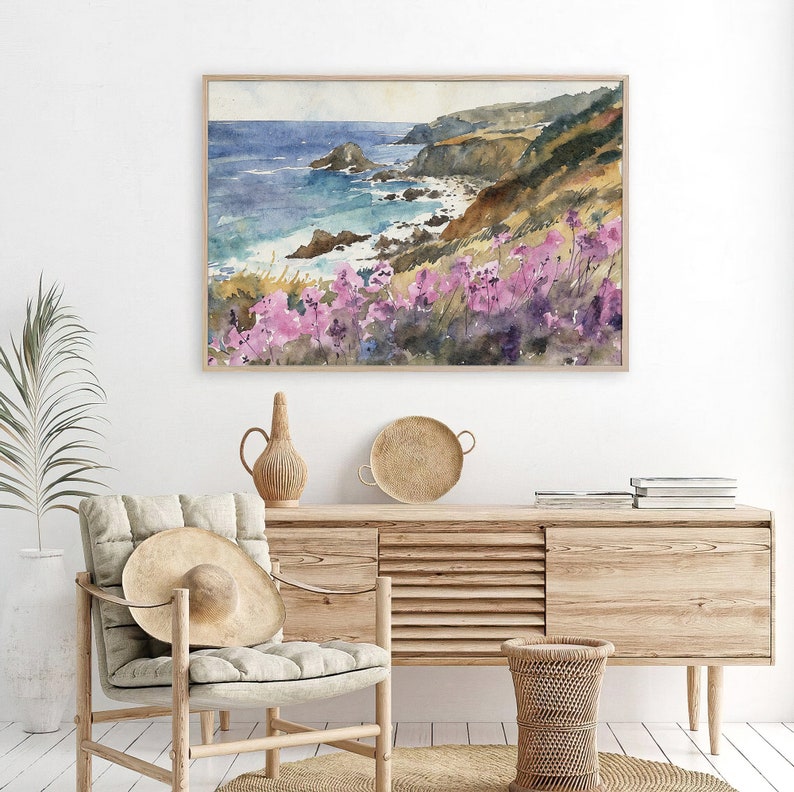 May include: A watercolor painting of a coastal scene with a rocky shoreline, blue ocean, and pink wildflowers in the foreground. The painting is framed in a light wood frame and is hanging on a white wall.