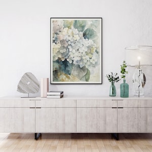 May include: A framed watercolor print of white and blue hydrangeas with green leaves. The artwork hangs above a white cabinet with decorative items, including a lamp, vases, and a stone sculpture. The room has a minimalist aesthetic.