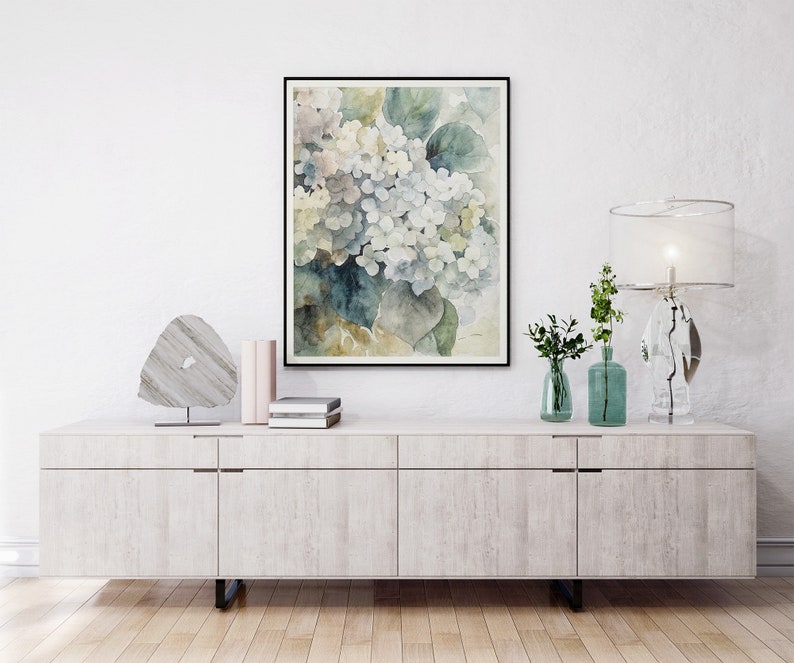 May include: A framed watercolor print of white and blue hydrangeas with green leaves. The artwork hangs above a white cabinet with decorative items, including a lamp, vases, and a stone sculpture. The room has a minimalist aesthetic.