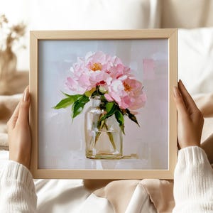 May include: A framed painting of pink peonies in a clear glass vase. The artwork features a light background and green leaves. The frame is a light wood color. The painting is held by a person wearing a white sweater.