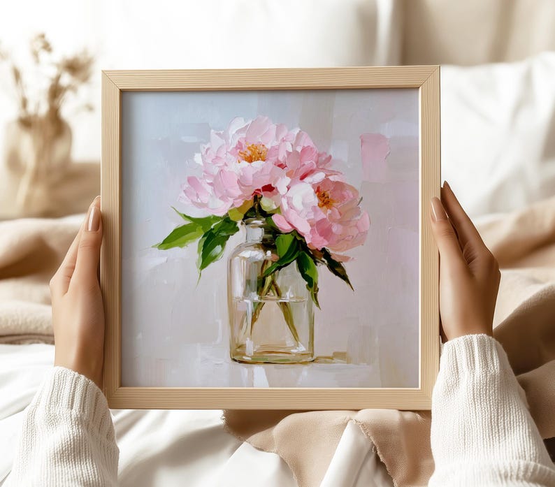 May include: A framed painting of pink peonies in a clear glass vase. The artwork features a light background and green leaves. The frame is a light wood color. The painting is held by a person wearing a white sweater.