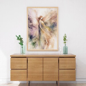 K&ouml;nnte beinhalten: Ein Aquarellbild einer Frau mit Fl&uuml;geln in Violett-, Gr&uuml;n- und Braunt&ouml;nen, gerahmt in einem hellen Holzrahmen, das &uuml;ber einem Holzschrank h&auml;ngt.