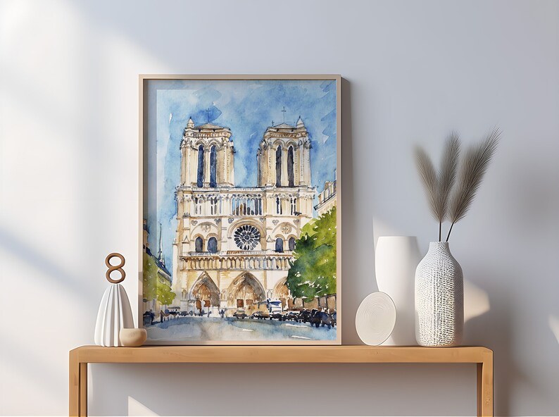 Op de afbeelding: Een aquarel schilderij van de Notre Dame kathedraal in Parijs, Frankrijk. Het schilderij is ingelijst en hangt aan een muur boven een houten consoletafel. De consoletafel heeft een witte vaas met gedroogd pampasgras en een wit keramiek beeldje erop.