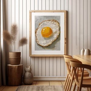 K&ouml;nnte beinhalten: Ein Gem&auml;lde eines Spiegeleis mit einem leuchtend gelben Eigelb auf wei&szlig;em Hintergrund. Das Ei ist in einem Holzrahmen gerahmt und h&auml;ngt an einer wei&szlig;en Wand.
