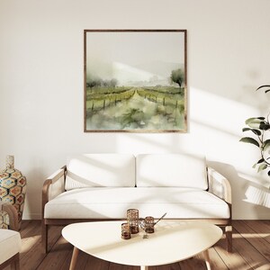 Op de afbeelding: Een witte bank met houten armleuningen en poten staat in een woonkamer met een witte muur en een ingelijst aquarel schilderij van een landschap met een pad dat naar een horizon leidt. Een plant in een pot staat in de hoek van de kamer.