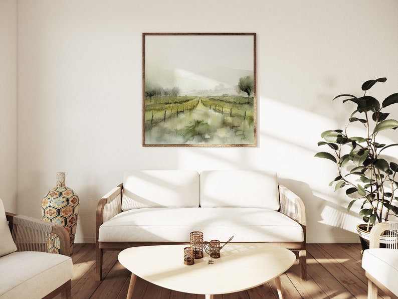 Op de afbeelding: Een witte bank met houten armleuningen en poten staat in een woonkamer met een witte muur en een ingelijst aquarel schilderij van een landschap met een pad dat naar een horizon leidt. Een plant in een pot staat in de hoek van de kamer.