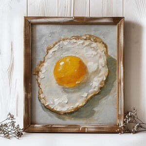 K&ouml;nnte beinhalten: Ein gerahmtes Gem&auml;lde eines Spiegeleis mit einem leuchtend gelben Eigelb. Das Ei ist in einem realistischen Stil mit sichtbaren Pinselstrichen gemalt. Der Rahmen ist ein einfacher Holzrahmen mit einer abgenutzten Oberfl&auml;che.