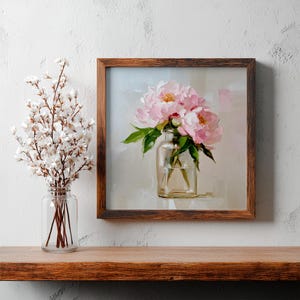 May include: A framed painting of pink peonies in a clear glass vase. The artwork has a light background and a dark brown wooden frame. A glass jar with white flowers is to the left of the painting.