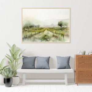 Op de afbeelding: Een aquarel van een wijngaardlandschap met een pad dat door de rijen wijnstokken loopt. Het schilderij is ingelijst in een licht houten lijst en hangt aan een witte muur boven een witte bank met grijze kussens.