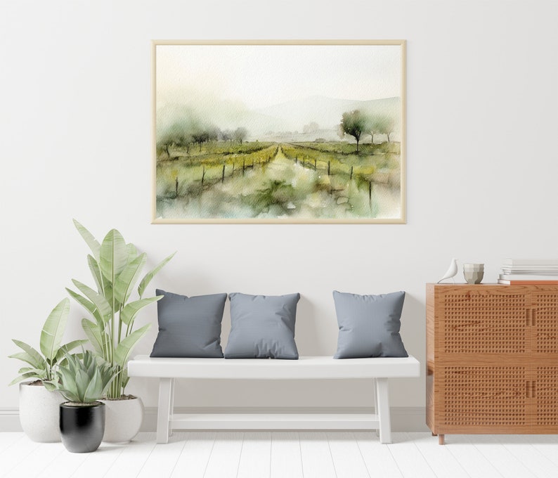 Op de afbeelding: Een aquarel van een wijngaardlandschap met een pad dat door de rijen wijnstokken loopt. Het schilderij is ingelijst in een licht houten lijst en hangt aan een witte muur boven een witte bank met grijze kussens.