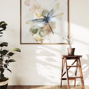 May include: Watercolor painting of a dragonfly with blue and yellow wings, set against a backdrop of soft green and beige foliage. The artwork is framed in a warm, wooden frame. The image is displayed on a white wall, with a wooden floor and a potted plant.