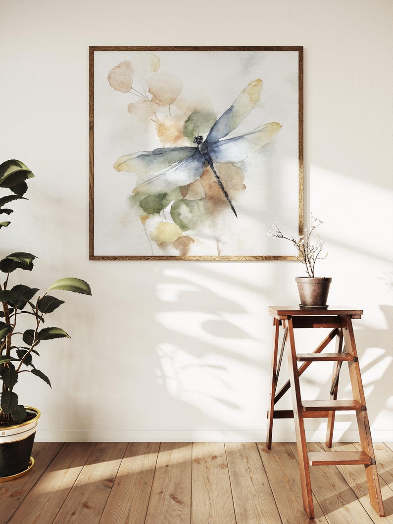 May include: Watercolor painting of a dragonfly with blue and yellow wings, set against a backdrop of soft green and beige foliage. The artwork is framed in a warm, wooden frame. The image is displayed on a white wall, with a wooden floor and a potted plant.