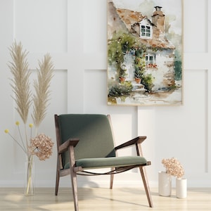 May include: A watercolor painting of a small cottage with a green roof and white walls. The cottage has a window with red flowers in the window box. The painting is framed in a light wood frame and is hanging on a white wall.