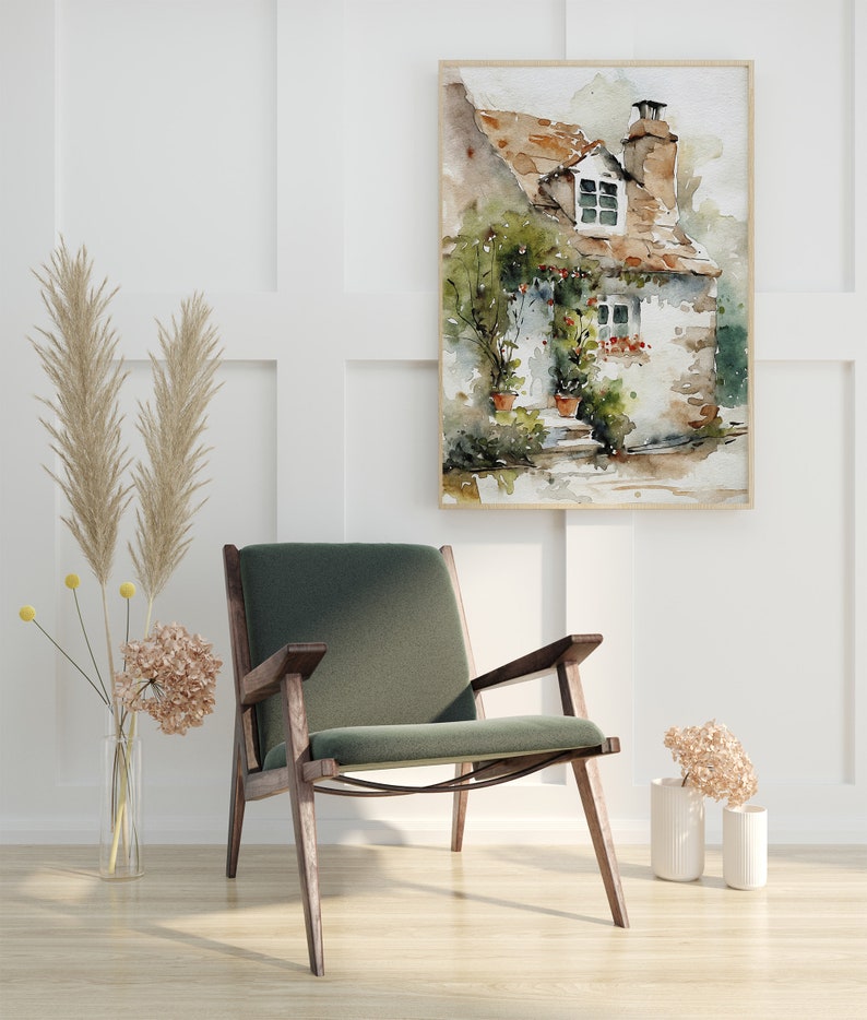 May include: A watercolor painting of a small cottage with a green roof and white walls. The cottage has a window with red flowers in the window box. The painting is framed in a light wood frame and is hanging on a white wall.