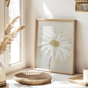May include: A framed painting of a white daisy with a yellow center on a light beige background. The frame is a light wood color. The painting is sitting on a table with a woven wicker tray and a white candle.