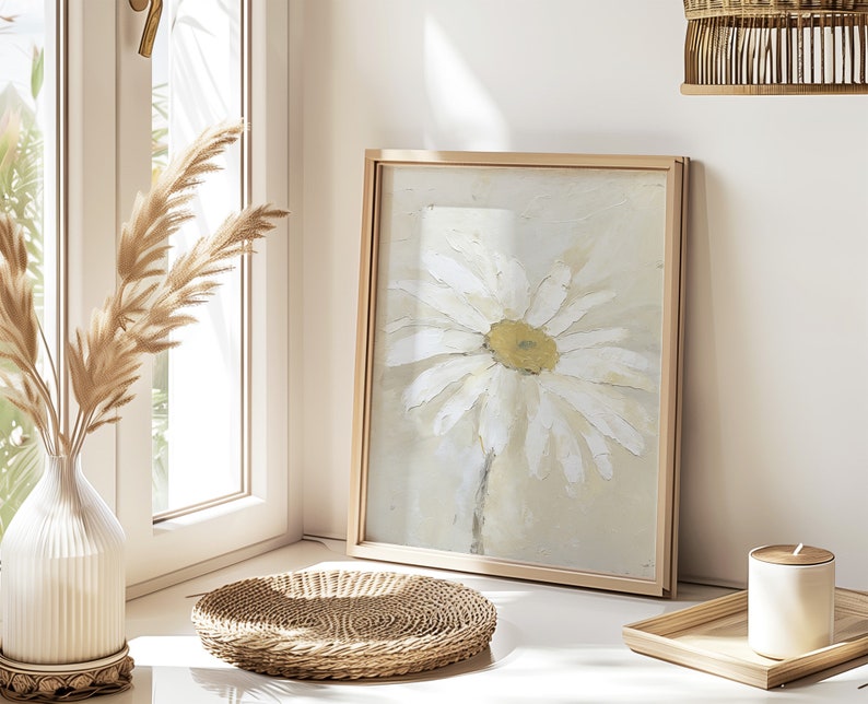 May include: A framed painting of a white daisy with a yellow center on a light beige background. The frame is a light wood color. The painting is sitting on a table with a woven wicker tray and a white candle.