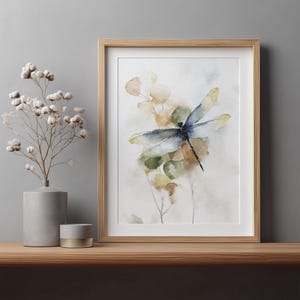 May include: Watercolor print of a dragonfly with blue and yellow wings, set against a backdrop of soft green and yellow foliage. The artwork is framed in a light wood frame and displayed on a wooden shelf.