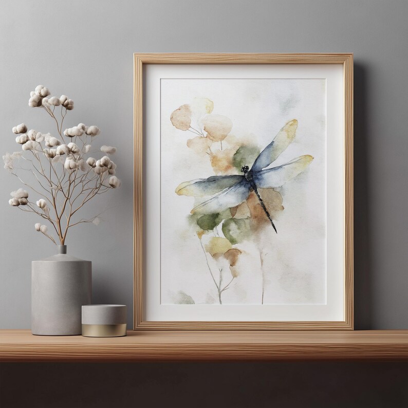 May include: Watercolor print of a dragonfly with blue and yellow wings, set against a backdrop of soft green and yellow foliage. The artwork is framed in a light wood frame and displayed on a wooden shelf.