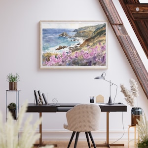 May include: A watercolor painting of a coastal scene with a rocky shoreline, blue ocean, and purple wildflowers in the foreground. The painting is framed and hanging on a white wall in a home office setting.