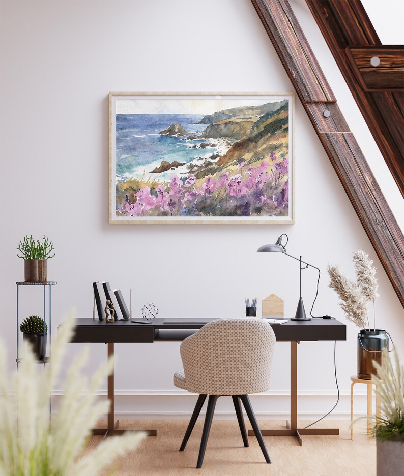 May include: A watercolor painting of a coastal scene with a rocky shoreline, blue ocean, and purple wildflowers in the foreground. The painting is framed and hanging on a white wall in a home office setting.