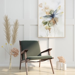 May include: An interior design scene featuring a framed watercolor painting of a dragonfly with a green and blue color scheme. A mid-century modern style chair with a green cushion and wooden frame sits below the artwork. Dried flowers in vases complete the decor.