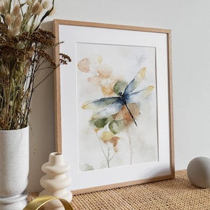 May include: Watercolor print of a dragonfly with blue and yellow wings, set against a backdrop of soft green, orange, and yellow foliage. The artwork is framed in a light brown wooden frame with a white mat.