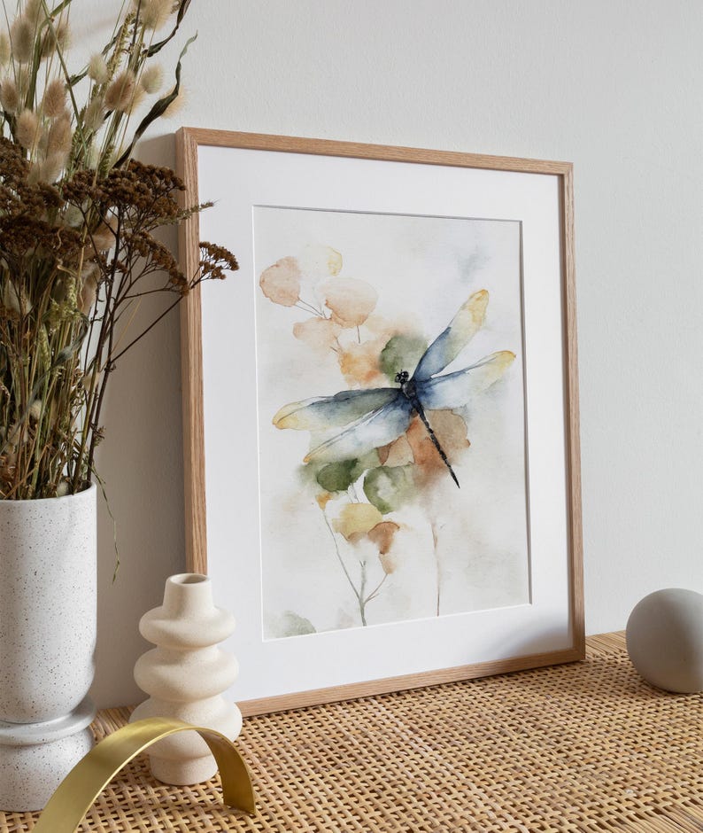 May include: Watercolor print of a dragonfly with blue and yellow wings, set against a backdrop of soft green, orange, and yellow foliage. The artwork is framed in a light brown wooden frame with a white mat.