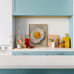 K&ouml;nnte beinhalten: Ein gerahmtes Gem&auml;lde eines Spiegeleis mit einem gelben Eigelb auf wei&szlig;em Hintergrund, das auf einer wei&szlig;en Arbeitsplatte mit einem blauen Schrank darunter ausgestellt ist. Auf der Arbeitsplatte befinden sich verschiedene K&uuml;chenutensilien, darunter eine rote Laterne, ein Glasgef&auml;&szlig; und Holzschneidebretter.