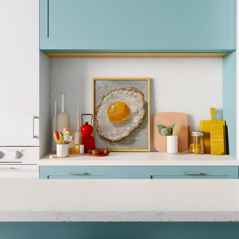 K&ouml;nnte beinhalten: Ein gerahmtes Gem&auml;lde eines Spiegeleis mit einem gelben Eigelb auf wei&szlig;em Hintergrund, das auf einer wei&szlig;en Arbeitsplatte mit einem blauen Schrank darunter ausgestellt ist. Auf der Arbeitsplatte befinden sich verschiedene K&uuml;chenutensilien, darunter eine rote Laterne, ein Glasgef&auml;&szlig; und Holzschneidebretter.