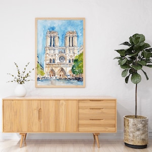 Op de afbeelding: Waterverf schilderij van de Notre Dame kathedraal in Parijs, Frankrijk. Het schilderij is ingelijst en hangt aan een witte muur boven een houten kast.