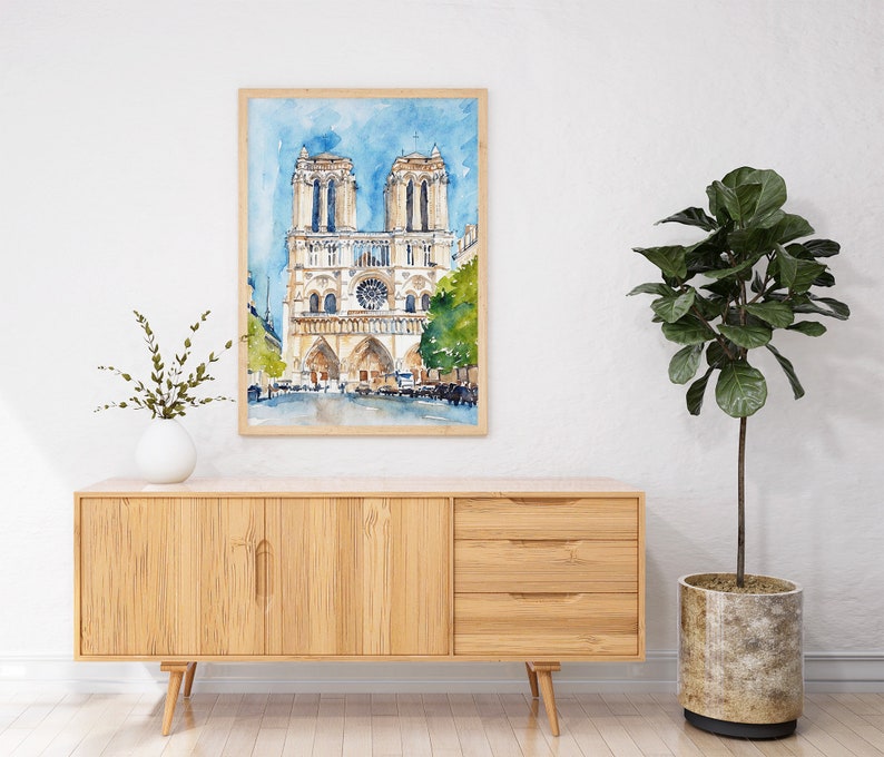 Op de afbeelding: Waterverf schilderij van de Notre Dame kathedraal in Parijs, Frankrijk. Het schilderij is ingelijst en hangt aan een witte muur boven een houten kast.