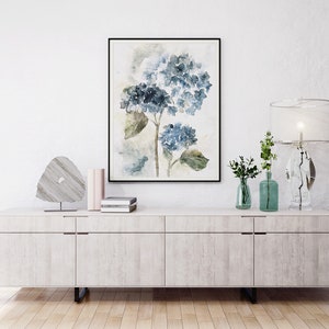 May include: A white dresser with four drawers and a framed watercolor painting of blue hydrangeas above it. The dresser has a light wood grain finish and silver drawer pulls. There are two small vases with green plants and a crystal lamp on the dresser.