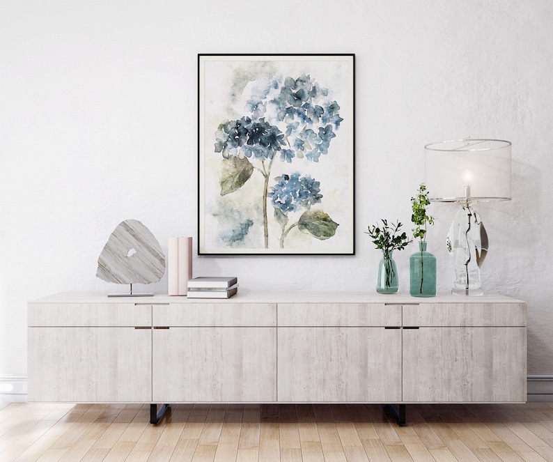 May include: A white dresser with four drawers and a framed watercolor painting of blue hydrangeas above it. The dresser has a light wood grain finish and silver drawer pulls. There are two small vases with green plants and a crystal lamp on the dresser.