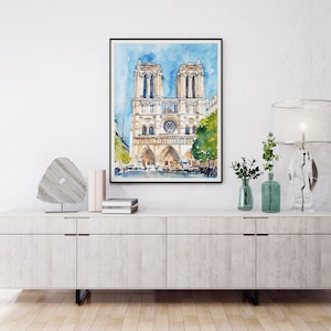 Op de afbeelding: Een aquarel van de Notre Dame kathedraal in Parijs, Frankrijk. Het schilderij is zwart ingelijst en hangt aan een witte muur boven een licht houten kast.