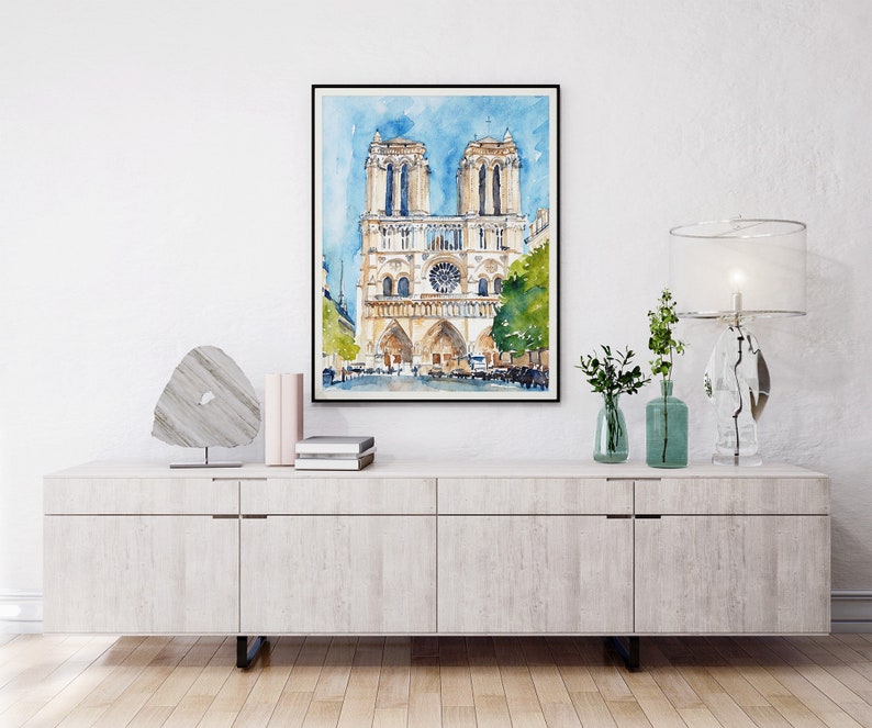 Op de afbeelding: Een aquarel van de Notre Dame kathedraal in Parijs, Frankrijk. Het schilderij is zwart ingelijst en hangt aan een witte muur boven een licht houten kast.