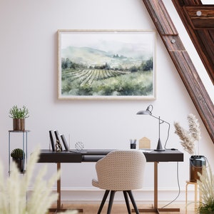 May include: A watercolor painting of a vineyard landscape with rolling hills, green vines, and a distant town. The painting is framed in a light wood frame and hangs on a white wall above a desk with a chair.