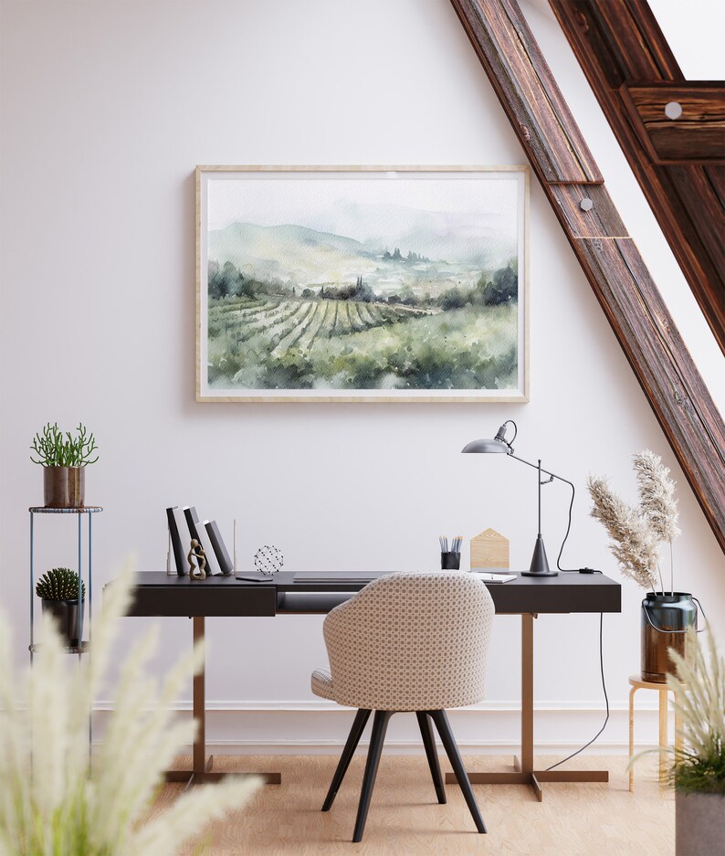 May include: A watercolor painting of a vineyard landscape with rolling hills, green vines, and a distant town. The painting is framed in a light wood frame and hangs on a white wall above a desk with a chair.