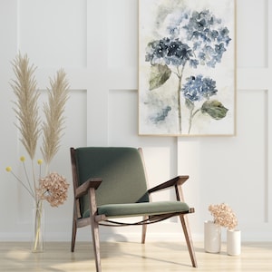 May include: A green velvet armchair with wooden legs sits in front of a white wall with a framed watercolor painting of blue hydrangeas.