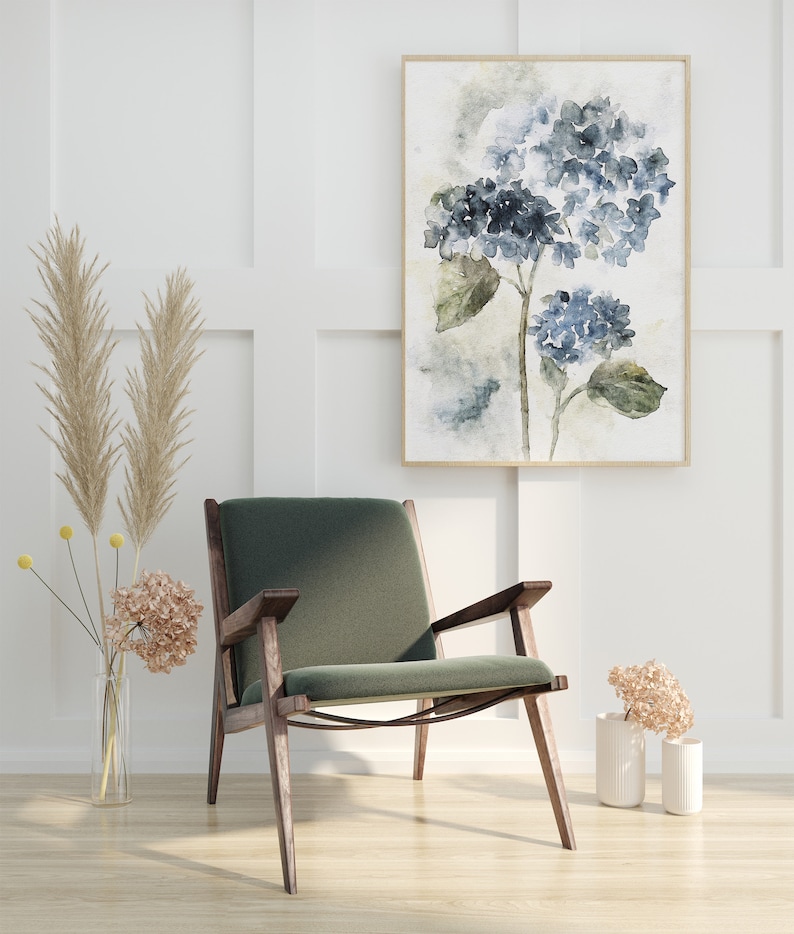 May include: A green velvet armchair with wooden legs sits in front of a white wall with a framed watercolor painting of blue hydrangeas.