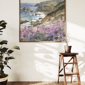 May include: A watercolor painting of a coastal landscape with a rocky shoreline, blue ocean, and purple wildflowers in the foreground. The painting is framed in a wooden frame.