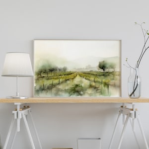 Op de afbeelding: Een aquarel van een wijngaardlandschap met een pad dat naar een verre horizon leidt. Het schilderij is ingelijst in een licht houten lijst en wordt tentoongesteld op een houten tafel met een witte lamp en een vaas met takken.