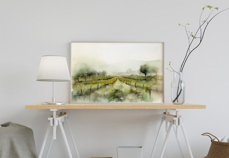 Op de afbeelding: Een aquarel van een wijngaardlandschap met een pad dat naar een verre horizon leidt. Het schilderij is ingelijst in een licht houten lijst en wordt tentoongesteld op een houten tafel met een witte lamp en een vaas met takken.