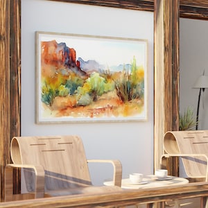 Puede incluir: Una pintura de acuarela de un paisaje des&eacute;rtico con formaciones rocosas rojas, arbustos verdes y un cielo azul. La pintura est&aacute; enmarcada en un marco de madera clara y est&aacute; colgada en una pared blanca.