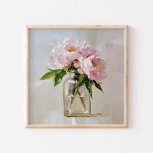 May include: A framed painting of pink peonies in a clear glass vase. The flowers have soft pink petals and yellow centers, with green leaves. The vase sits on a white surface against a neutral background. The frame is made of light-colored wood.