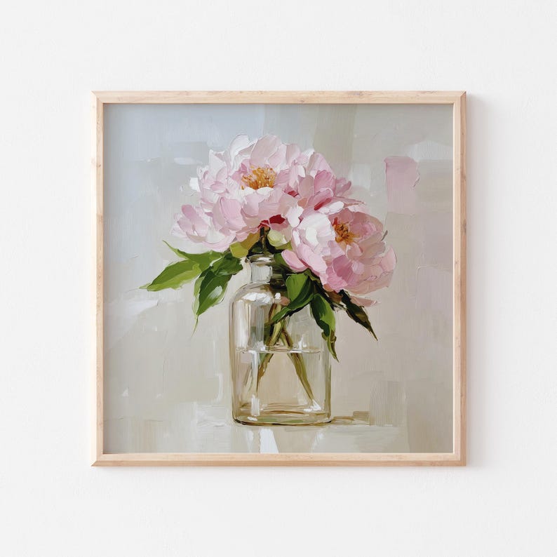 May include: A framed painting of pink peonies in a clear glass vase. The flowers have soft pink petals and yellow centers, with green leaves. The vase sits on a white surface against a neutral background. The frame is made of light-colored wood.