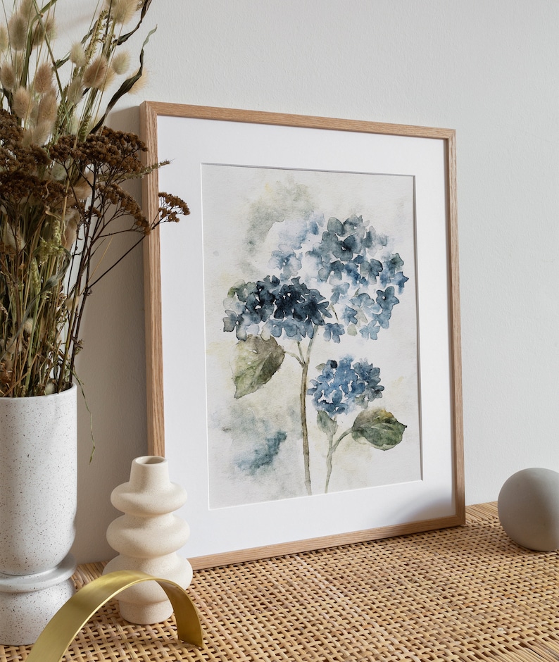 May include: A framed watercolor painting of blue hydrangeas with green leaves on a white background. The frame is made of light wood.